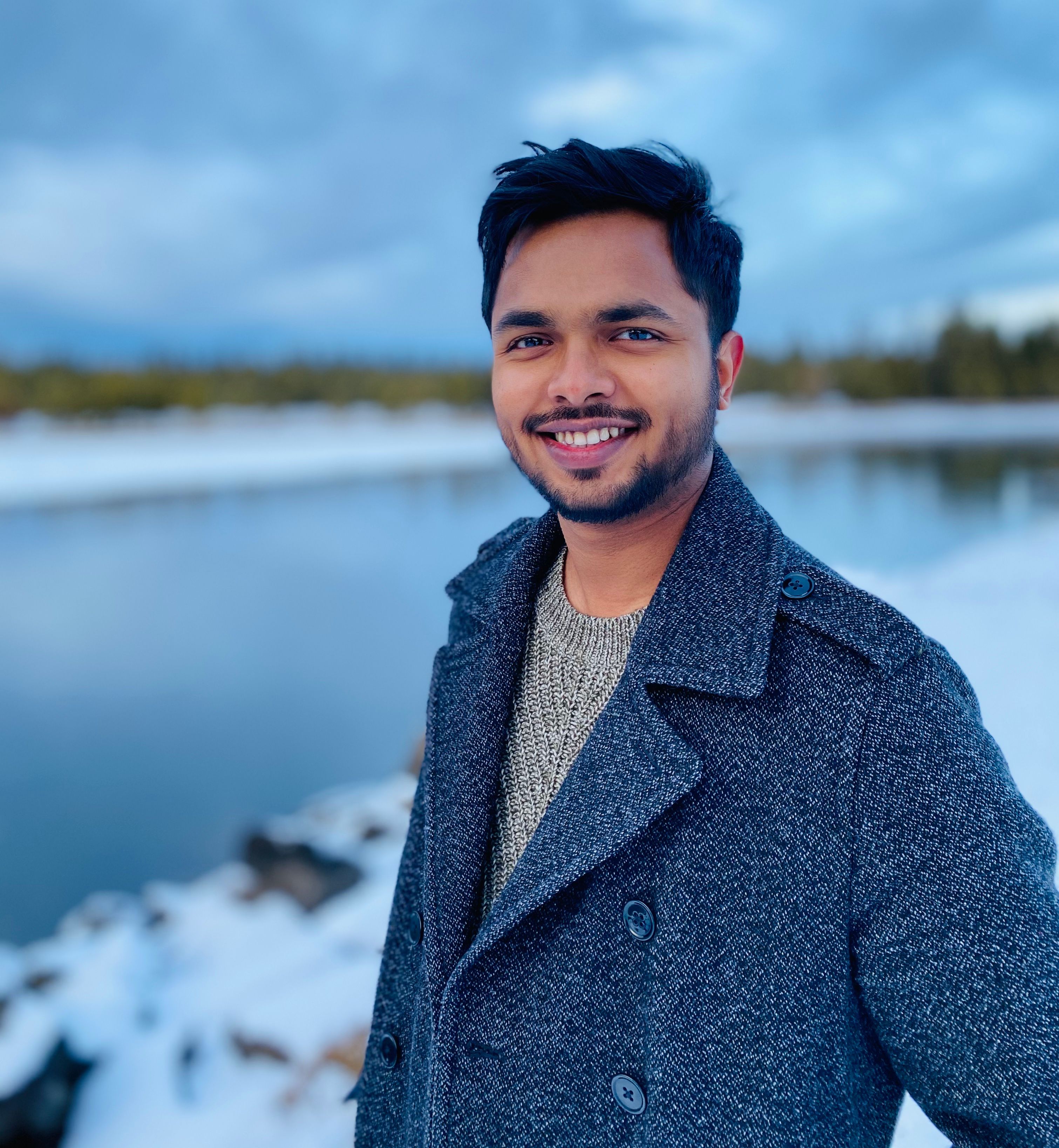 Proshonjit Mitra in a dark navy peacoat over a knit sweater, smiling, in front of a stormy blue-gray sky over a frozen lake and snow-covered shoreline.