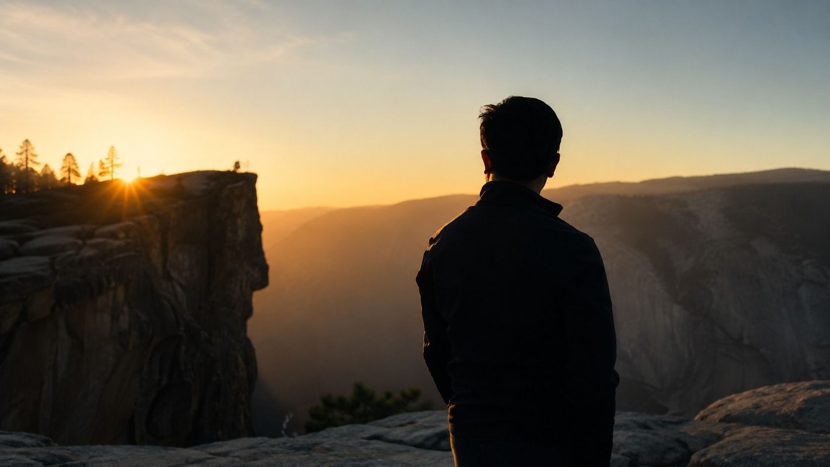 A person stands at the edge of a cliff at Yosemite, silhouetted against a warm sunrise, looking out across a wide canyon toward the horizon.